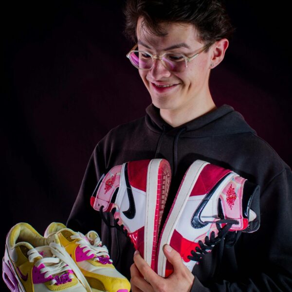 A young man joyfully displays two pairs of vibrant sneakers against a dark backdrop.